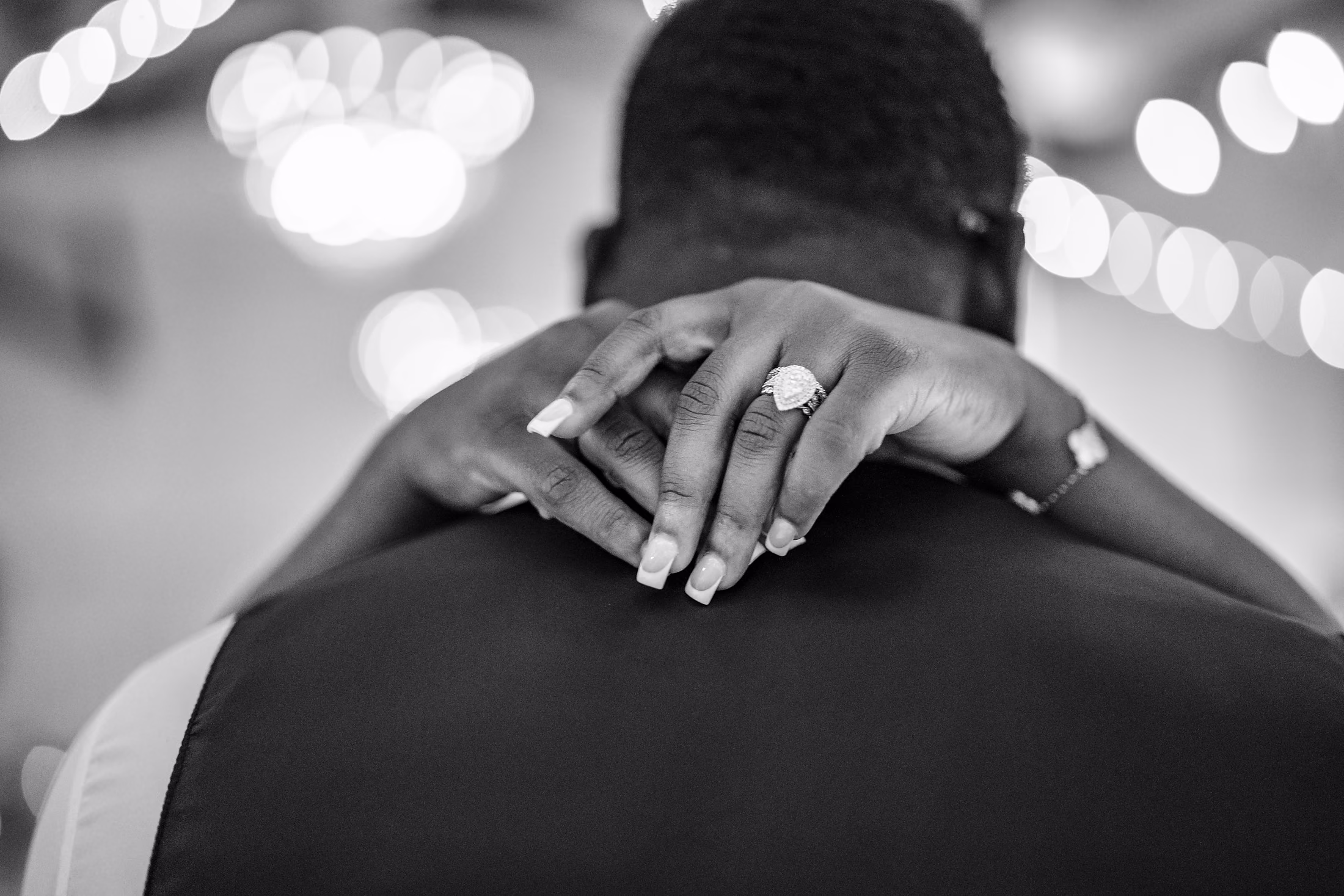 Black-and-white detail of a bride's ringed hand on her groom's neck