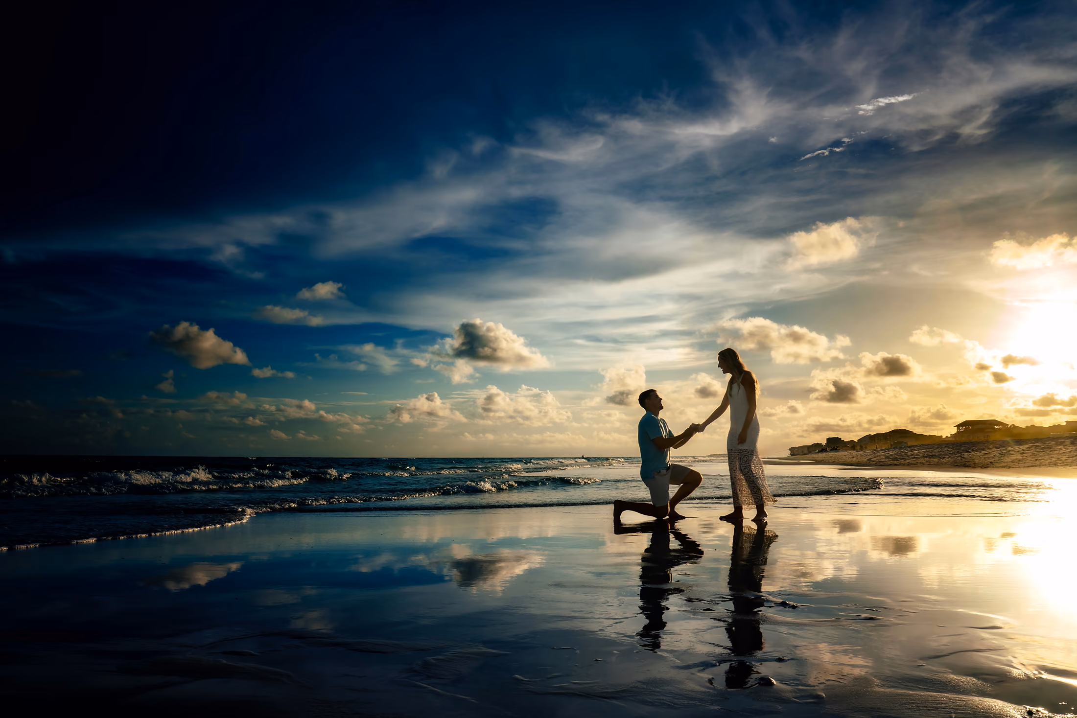 Couple at the water's edge at sunset, one kneeling on the wet sand
