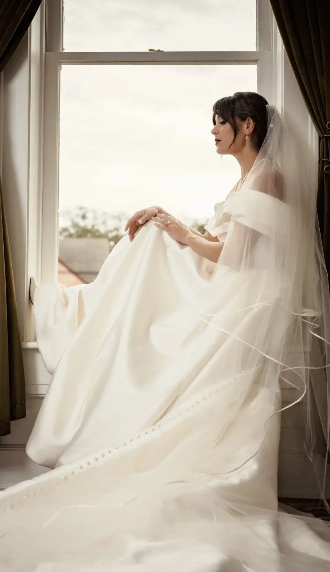 Bride backlit at the window, gown pooling around her